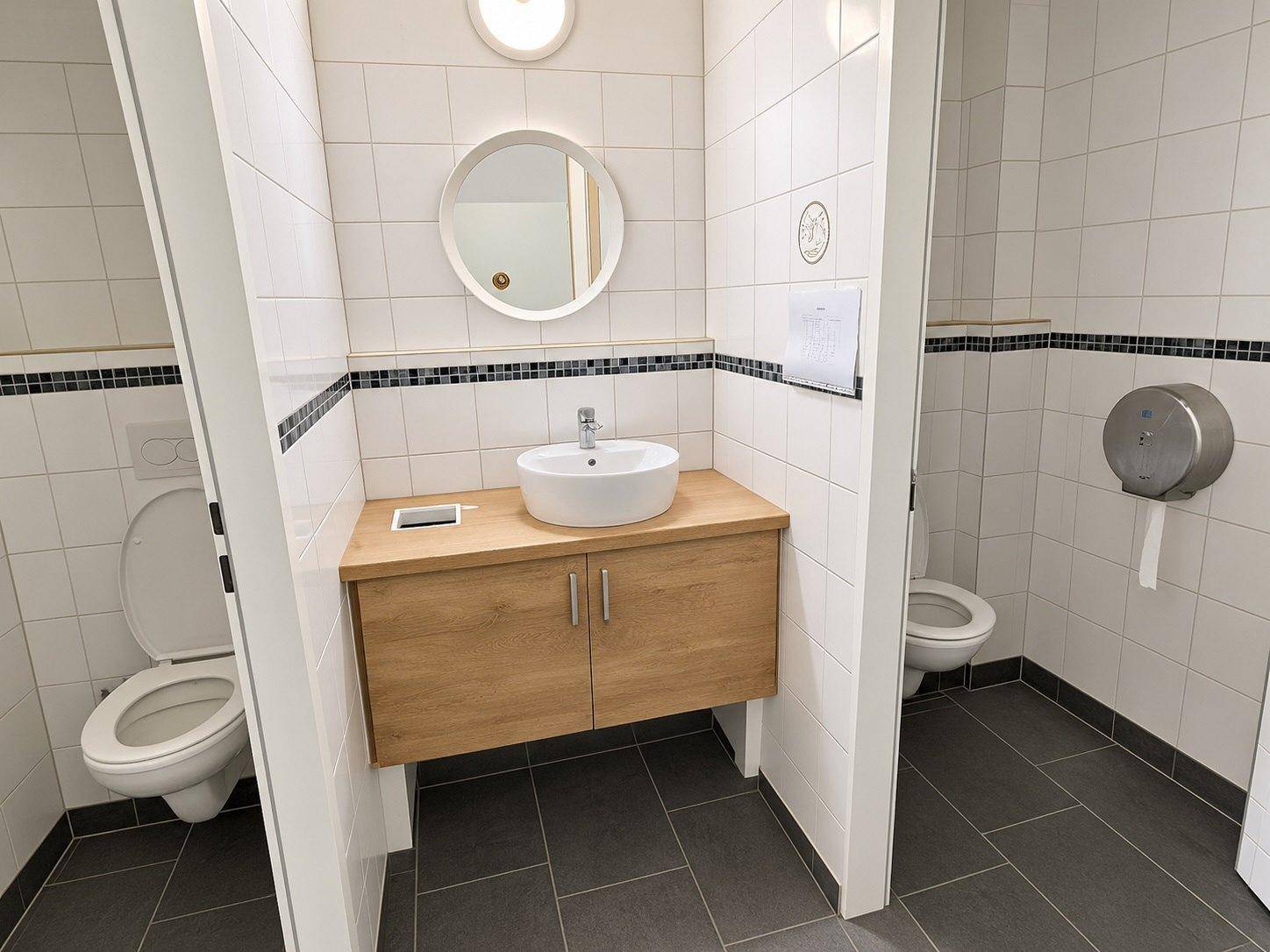 Moderne Toilettenanlage mit zwei Kabinen, Waschbecken und weißen Fliesen.
