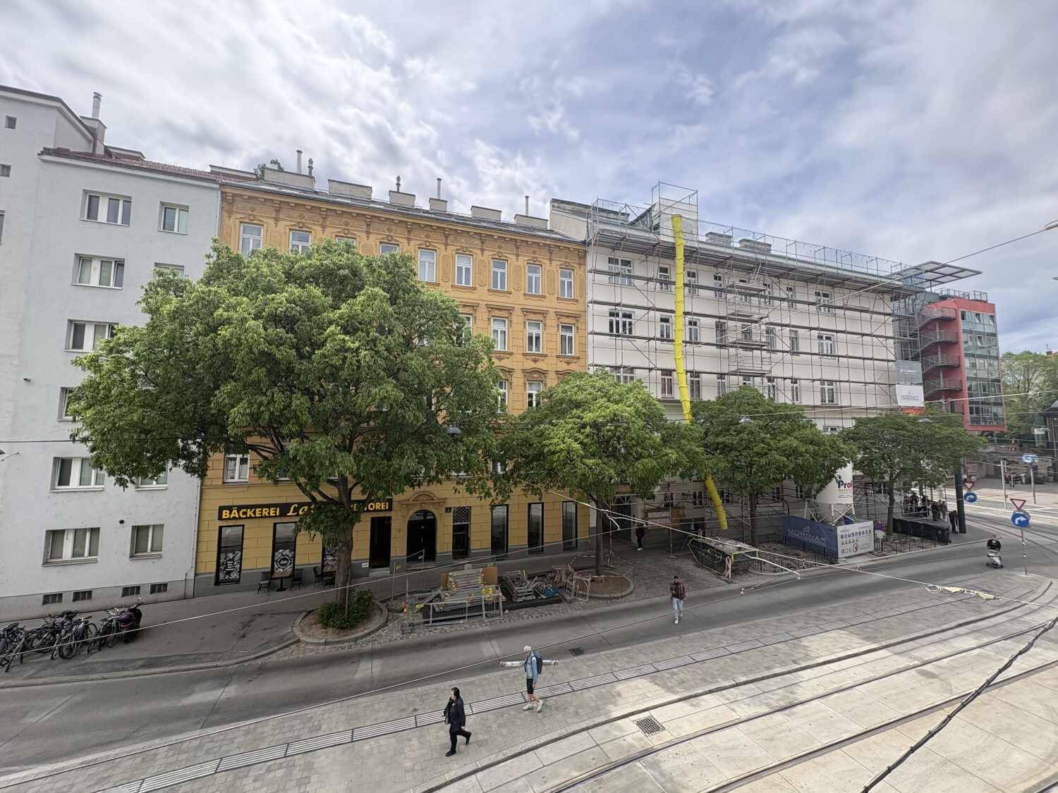 Straßenansicht mit Gebäuden, Bäumen und einer Baustelle mit Gerüst, die auf Renovierungsarbeiten hindeutet.