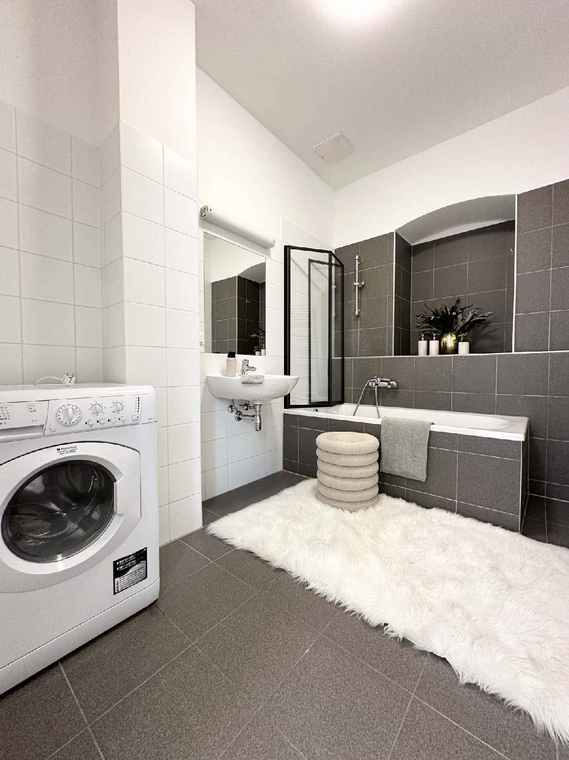 Modernes Badezimmer mit Badewanne, Dusche und Waschmaschine, ausgestattet mit grauen und weißen Fliesen.