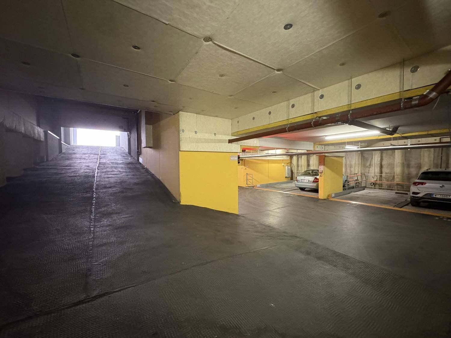Zufahrtsrampe zu einer Tiefgarage mit hellen Wänden und weiteren Parkplätzen im Hintergrund.