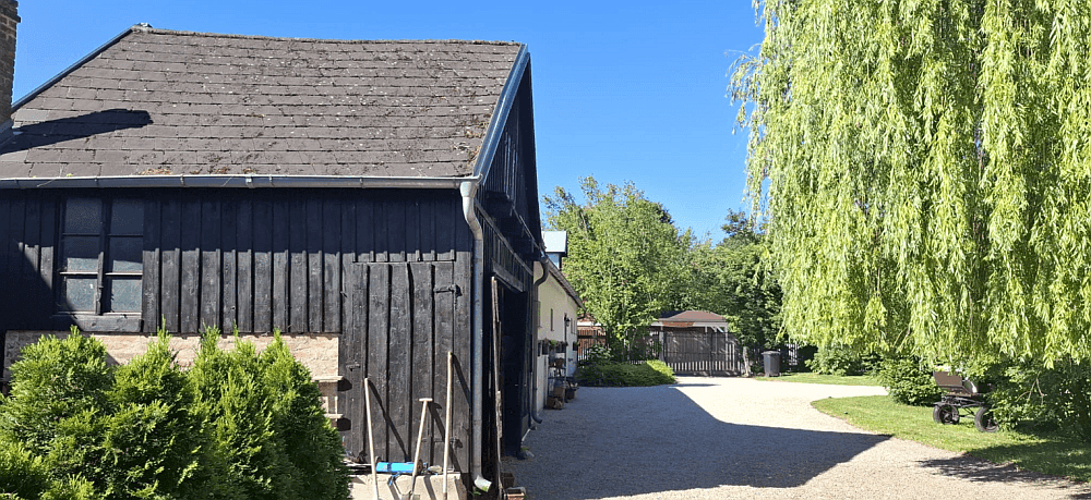 Rustikales Holzgebäude mit dunklem Dach und gepflegten Büschen entlang eines Kiesweges.