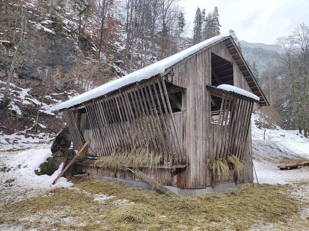 Rustikaler Holzschuppen oder Futterstelle mit Heu und Schnee in einer bergigen Umgebung.