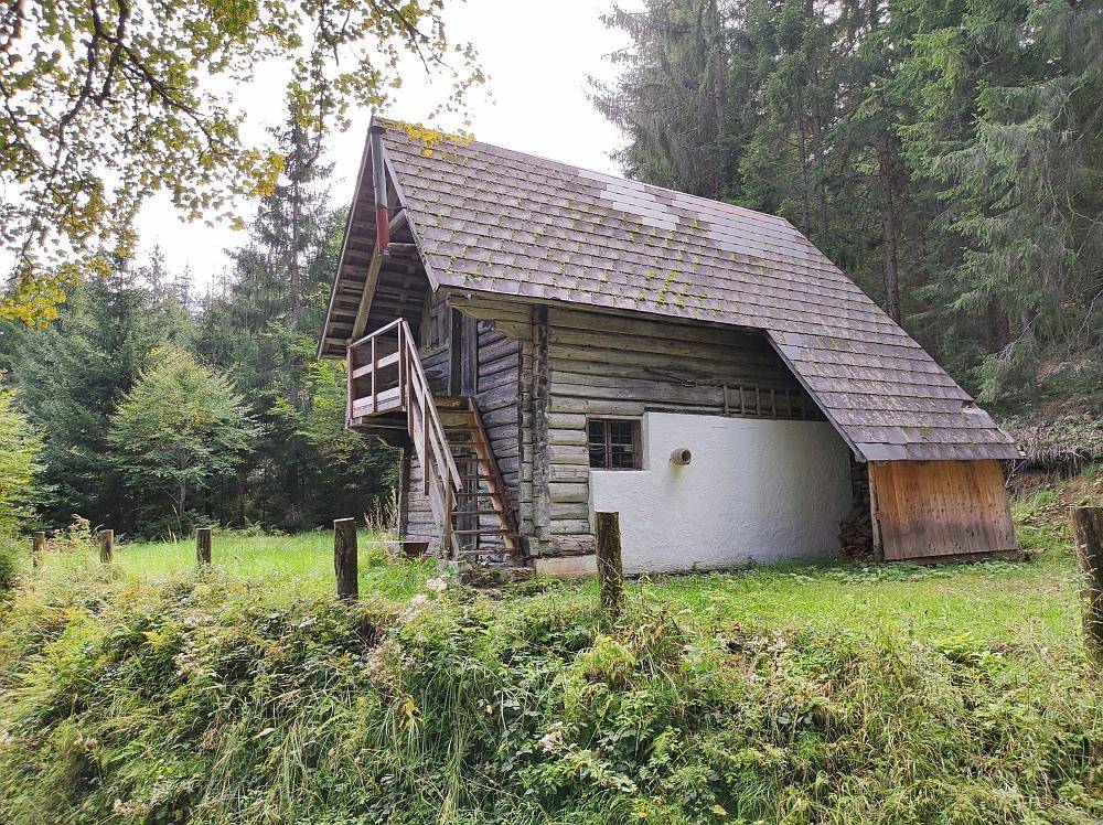 Rustikale Holzhütte mit Schindeldach und Außentreppe, umgeben von Wald und Wiese.