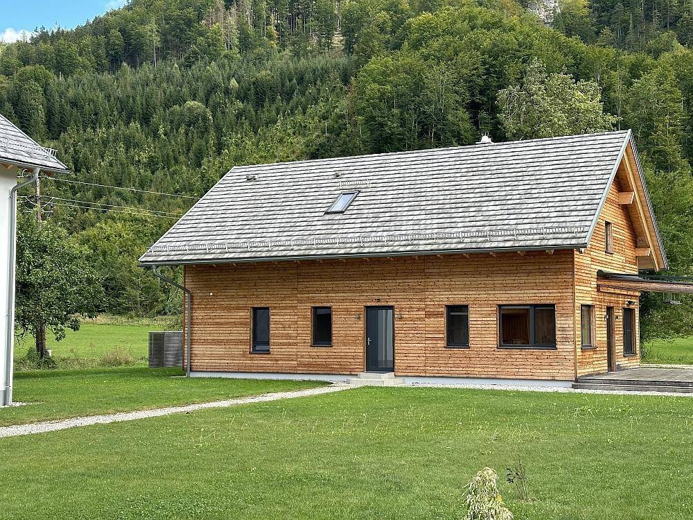 Modernes Holzhaus mit Schindeldach und großen Fenstern, umgeben von einer grünen Wiese.