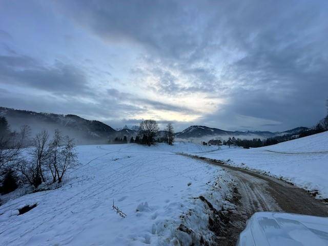Verschneite Landstraße führt durch ein Tal mit bewaldeten Bergen unter einem dramatischen Himmel.