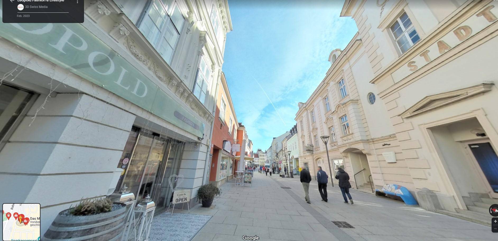 Belebte Straßenansicht einer historischen Gasse mit Gebäuden und Geschäften unter einem klaren blauen Himmel.