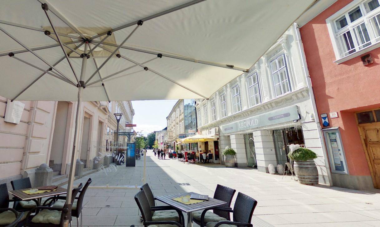Belebte Fußgängerzone mit Straßencafés und historischen Gebäuden unter einem großen Sonnenschirm.