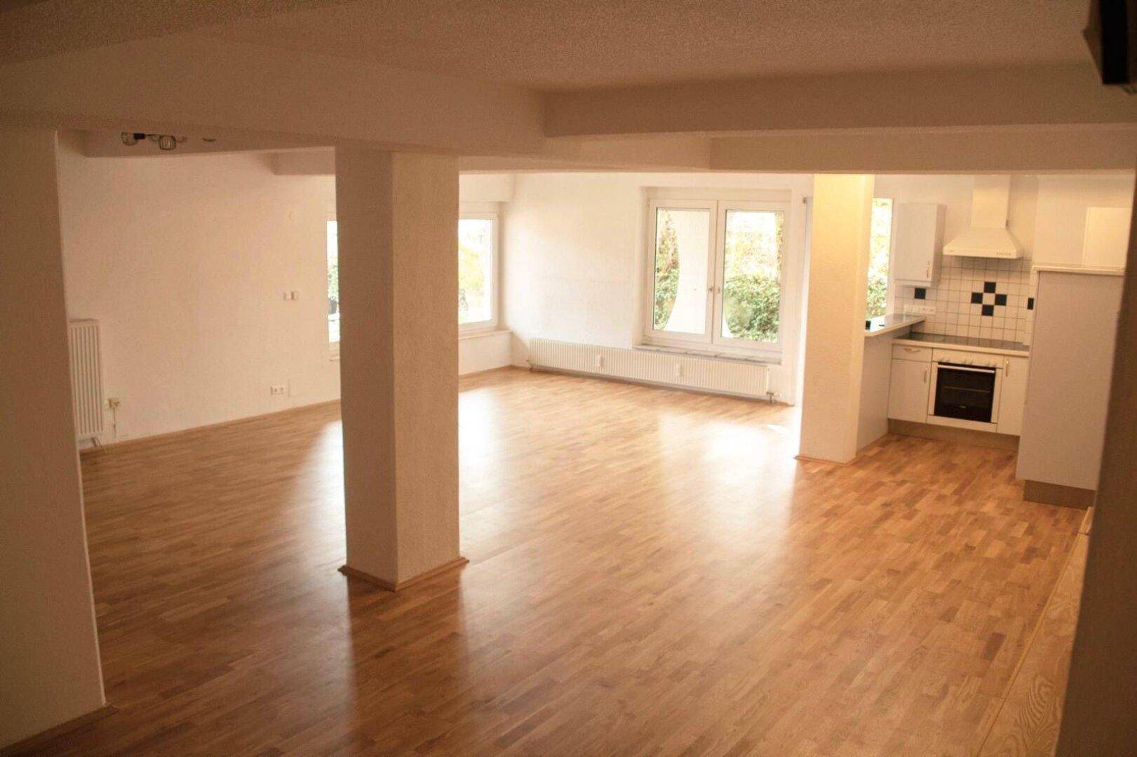 Geräumiger offener Wohnbereich mit Holzboden, großen Fenstern und integrierter Küchenzeile.