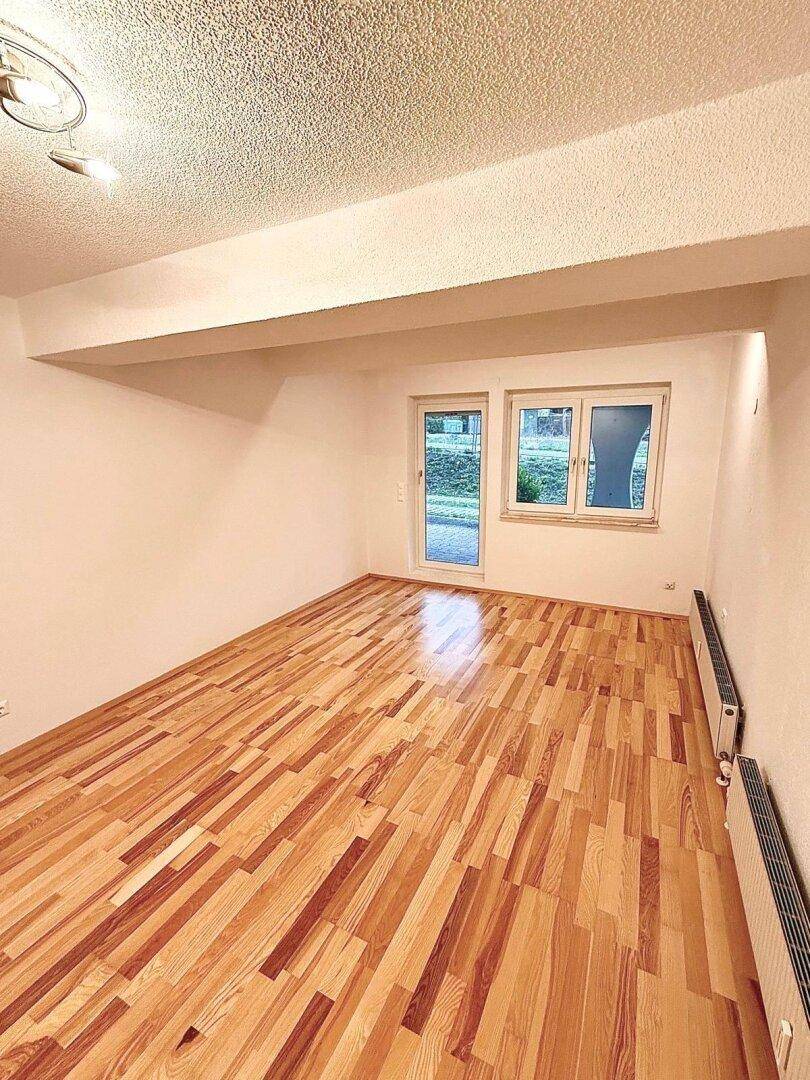 Helles Zimmer mit Holzboden, Zugang nach draußen und Heizkörpern unter den Fenstern.