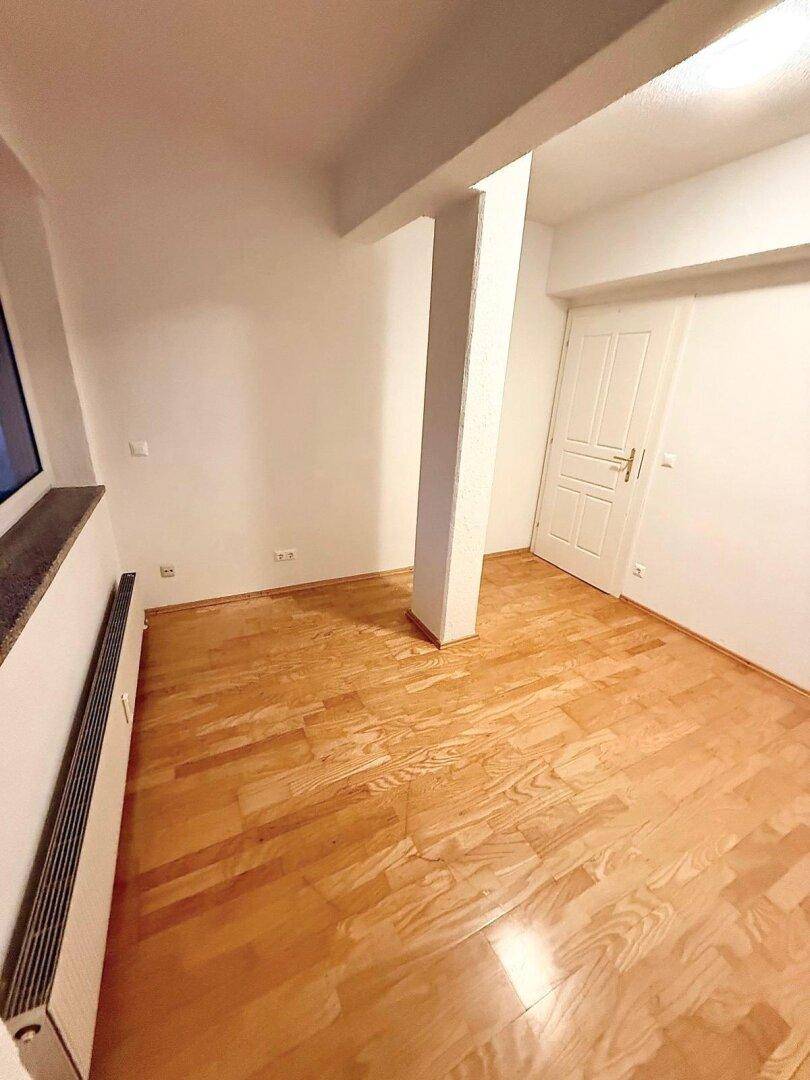 Leeres Zimmer mit Holzboden, einem tragenden Pfeiler und einem Heizkörper unter dem Fenster.