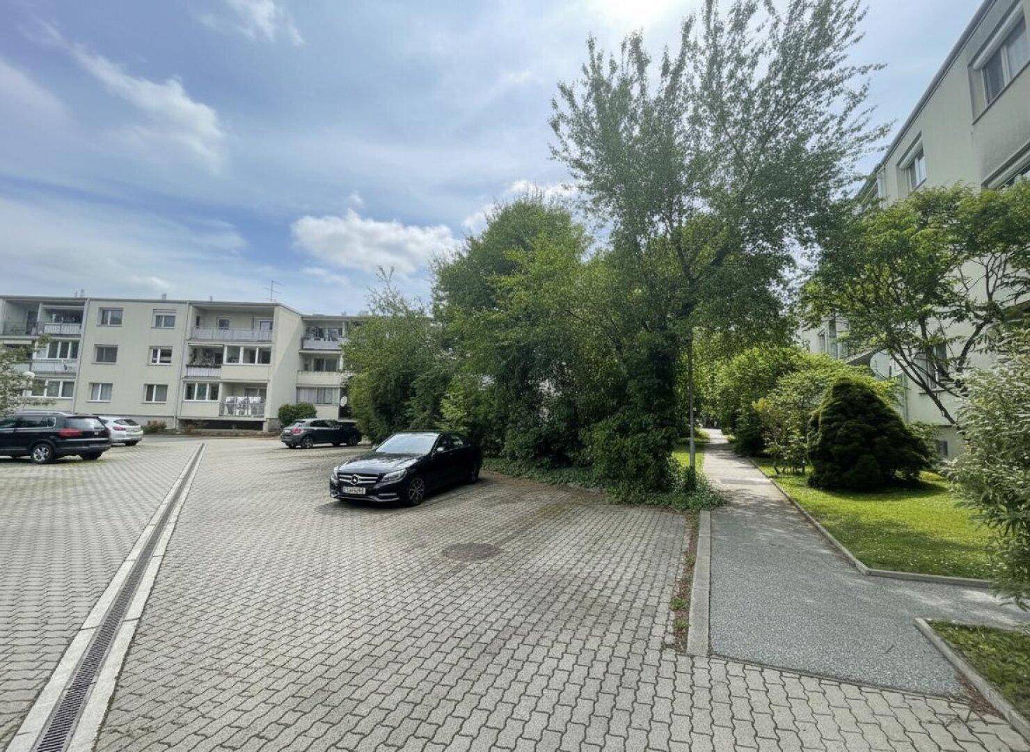Außenansicht des Gebäudes mit gepflastertem Parkplatz und viel Grünfläche an einem sonnigen Tag.