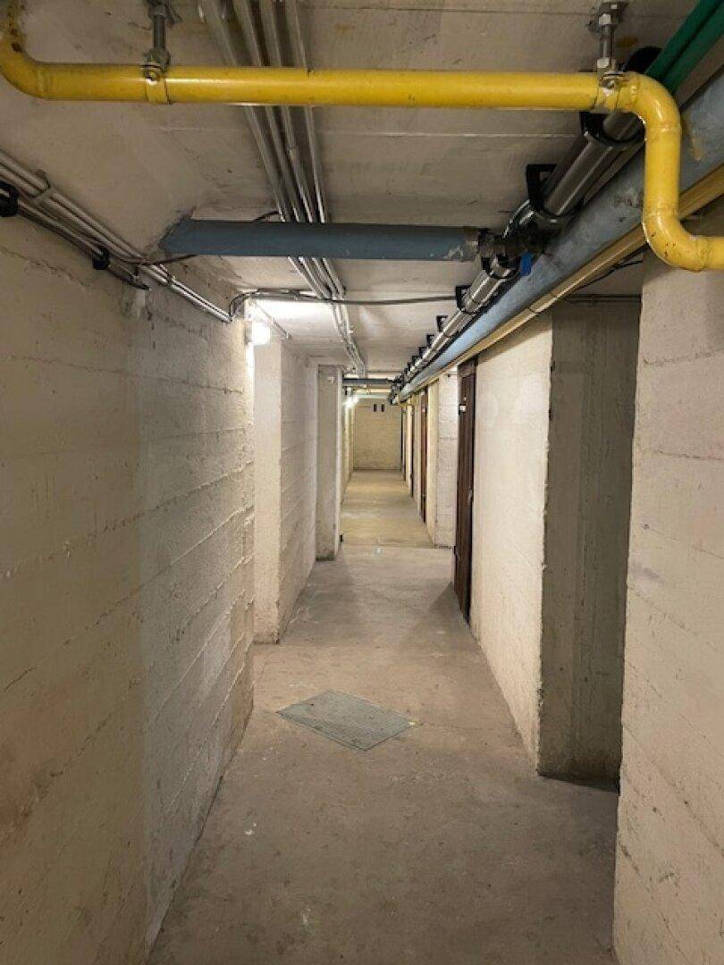 Langer Kellerflur mit Betonwänden, sichtbaren Rohren und Zugang zu Lagerräumen.