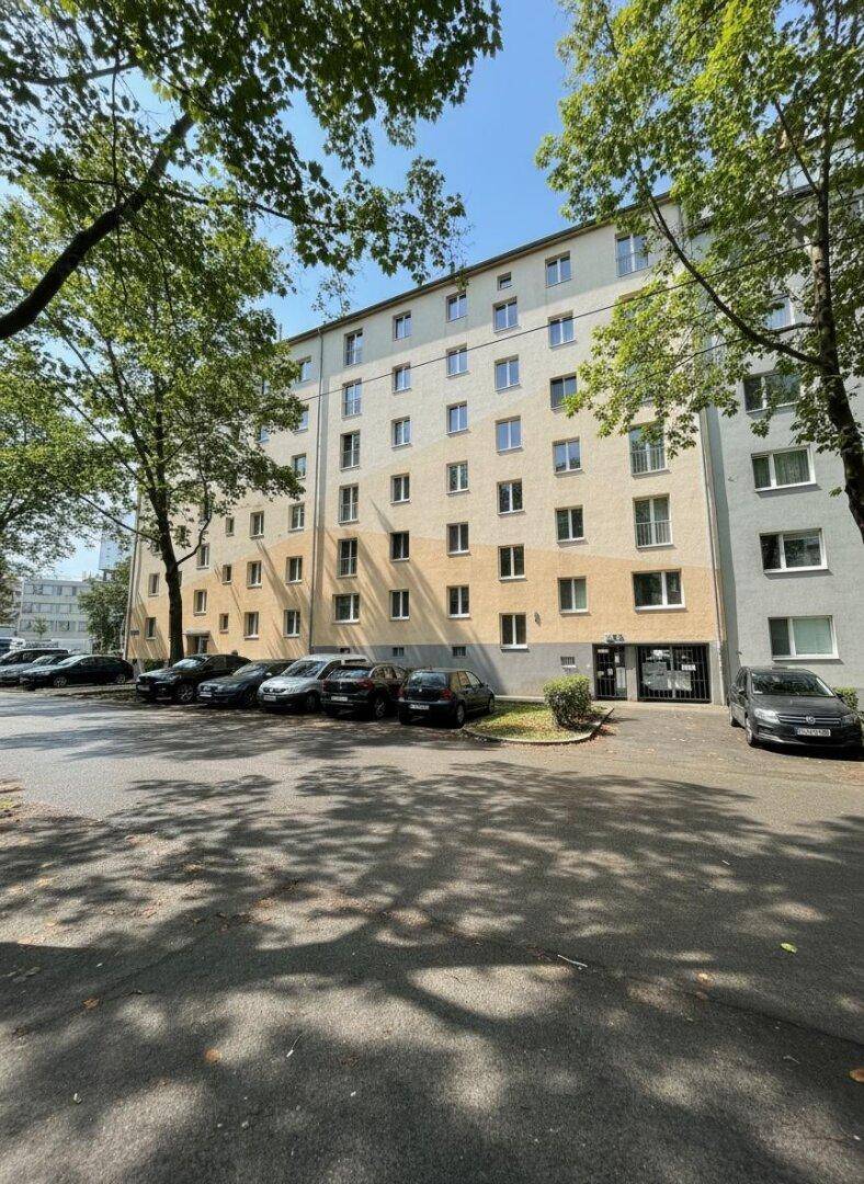 Mehrstöckiges Wohngebäude mit Parkplätzen davor, umgeben von Bäumen an einem sonnigen Tag.