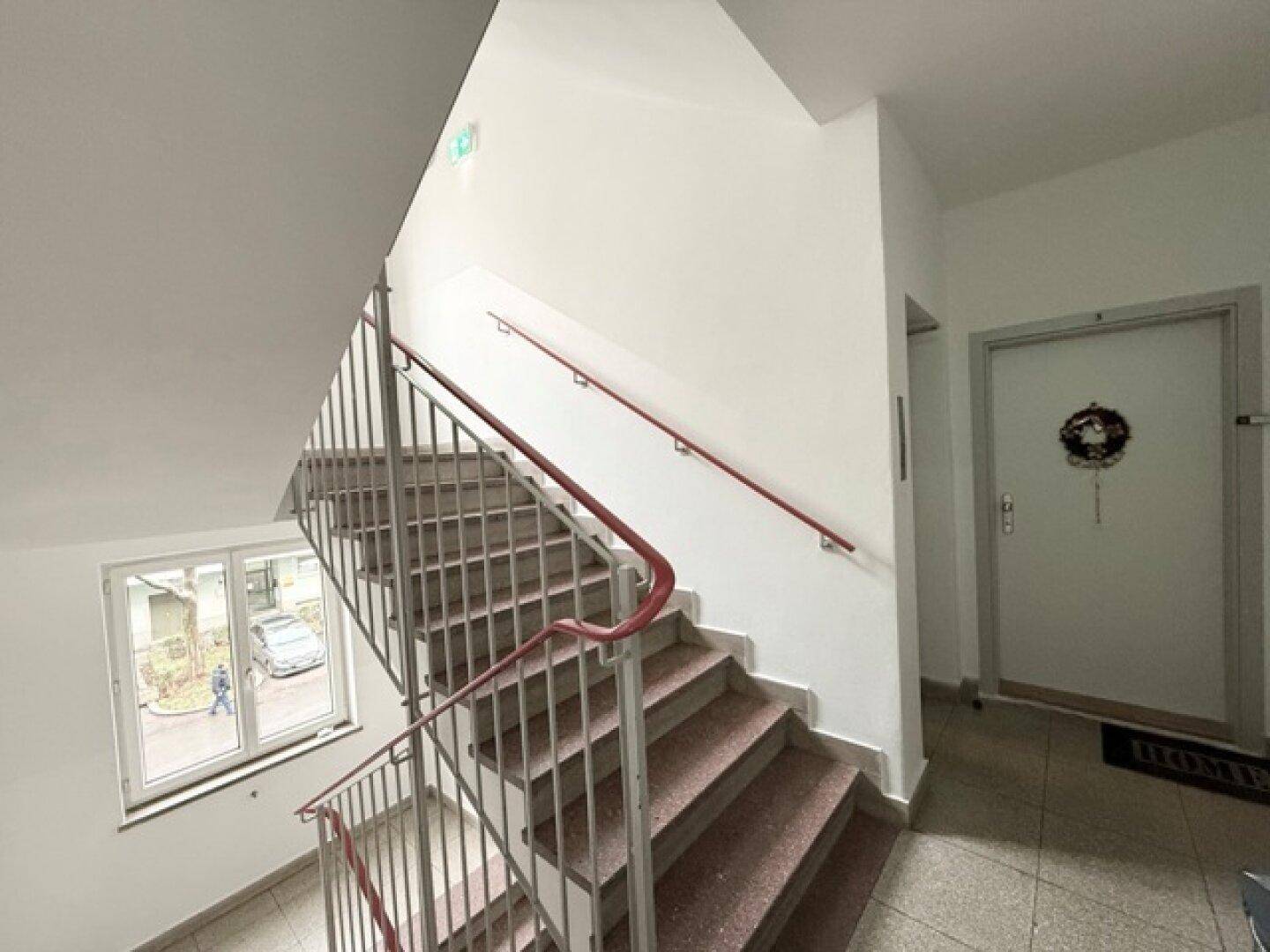 Heller Treppenaufgang in einem Wohngebäude mit rotem Handlauf, Fliesenboden und Fenster zum Außenbereich.