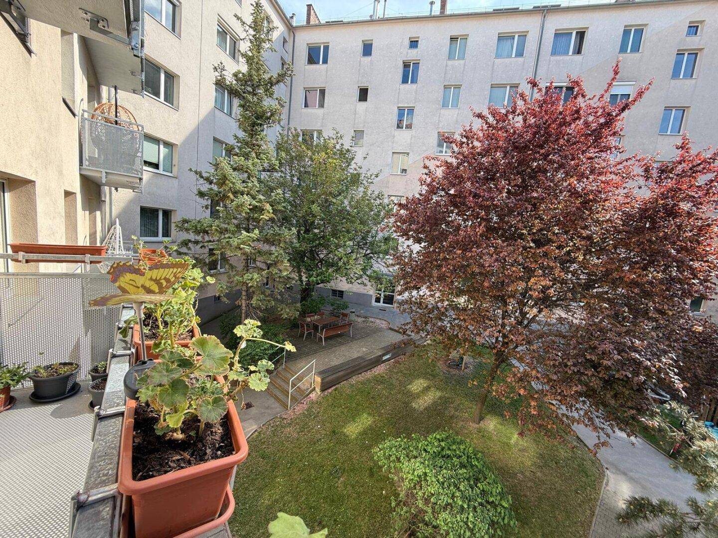 Blick vom Balkon auf einen gepflegten grünen Innenhof mit Bäumen und Sitzgelegenheiten.