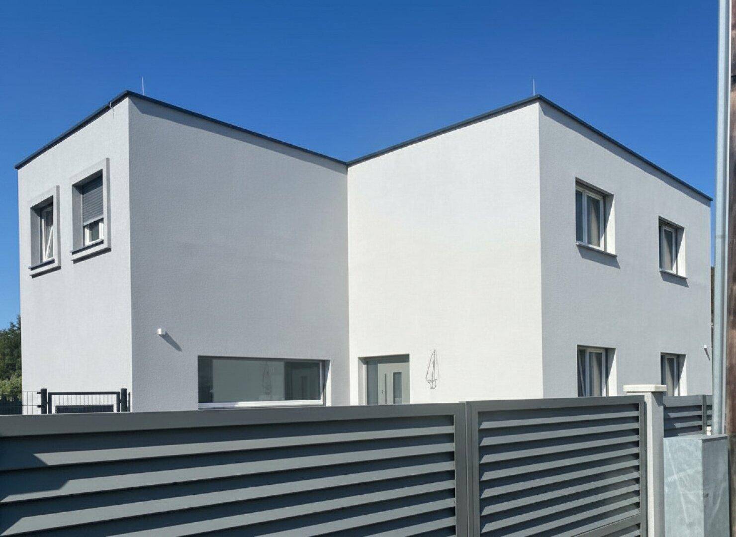 Zeitgenössisches Haus mit schlichter Fassade, umgeben von einem modernen Zaun und klarem Himmel.