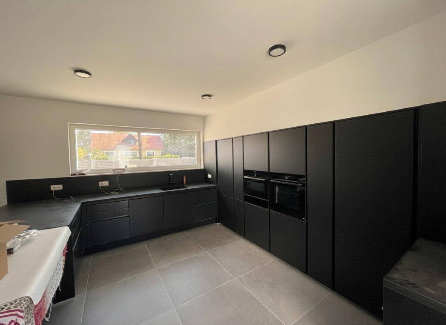Moderne schwarze Einbauküche mit integrierten Geräten und großem Fenster, noch ungenutzt.