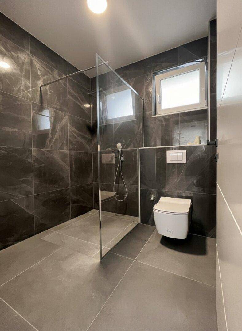 Modernes Badezimmer mit großformatigen dunklen Fliesen, ebenerdiger Dusche und Wand-WC.