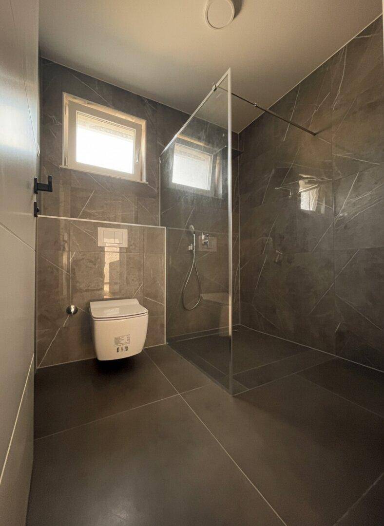 Elegantes Badezimmer mit dunklen Fliesen, geräumiger Dusche und modernem Wand-WC.