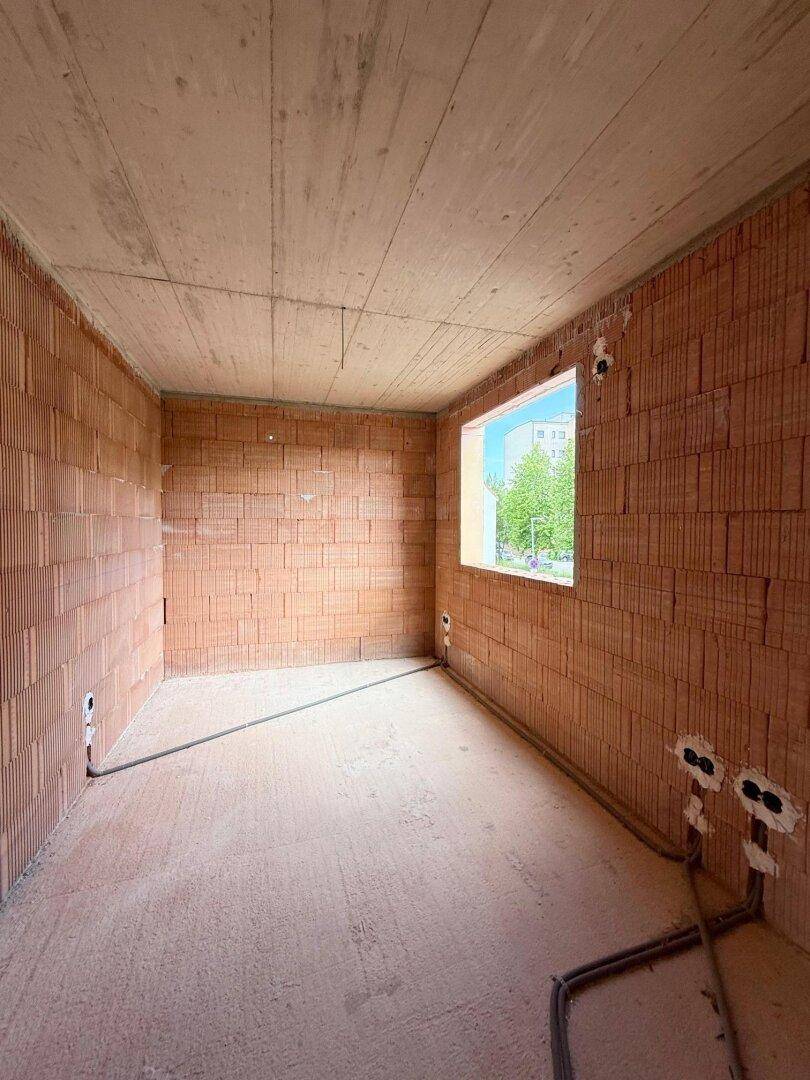 Zimmer im Rohbau mit einem Fenster und vorbereiteten Anschlüssen an den Wänden.