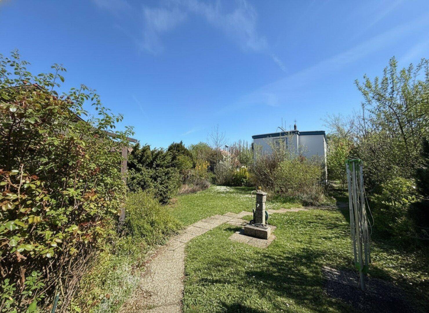 Sonniger Garten mit Steinpfad, dekorativem Element und einem Schuppen im Hintergrund.