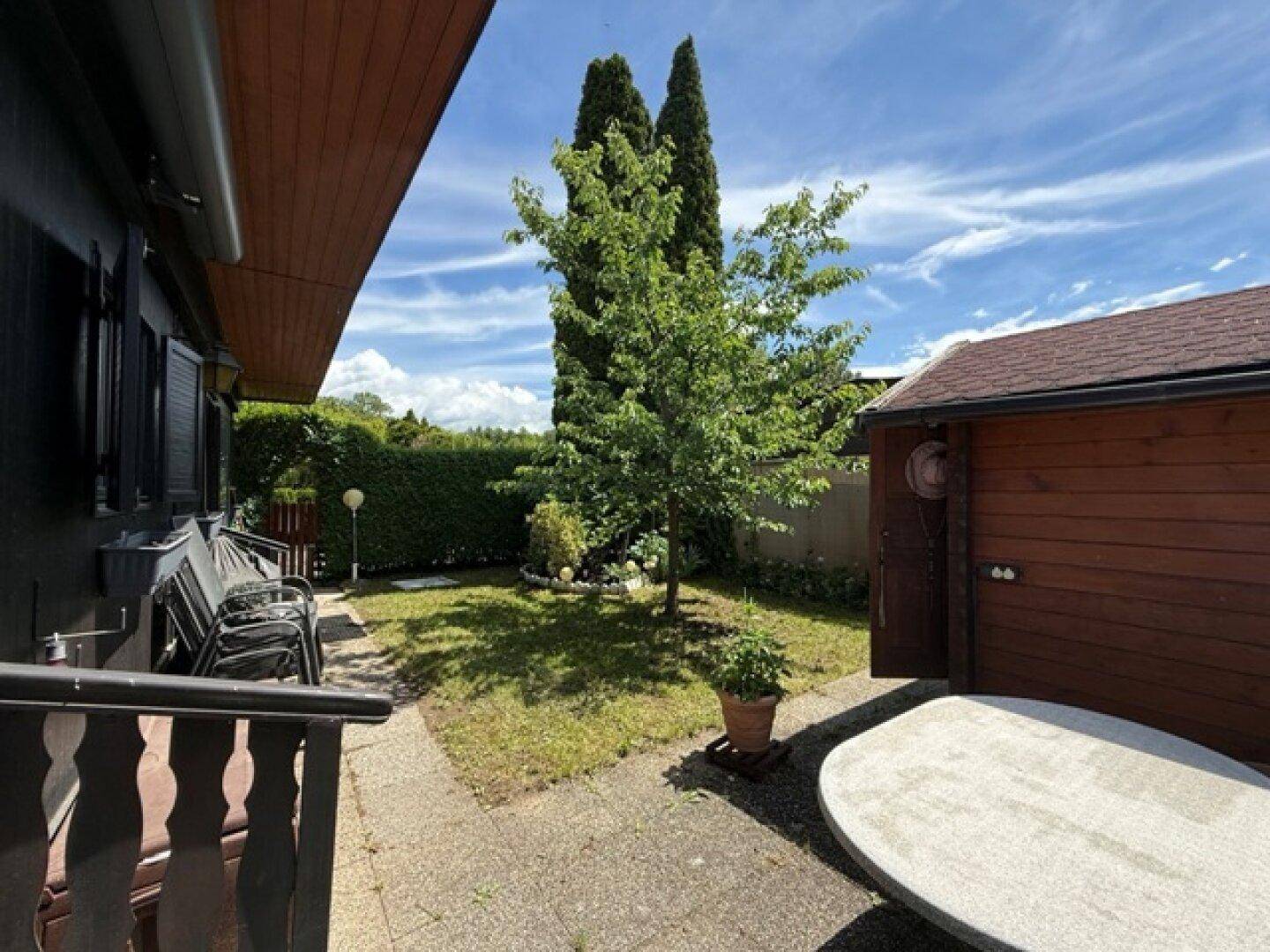 Gartenbereich mit Rasen, Baum, Gartenhaus und einem Steintisch.
