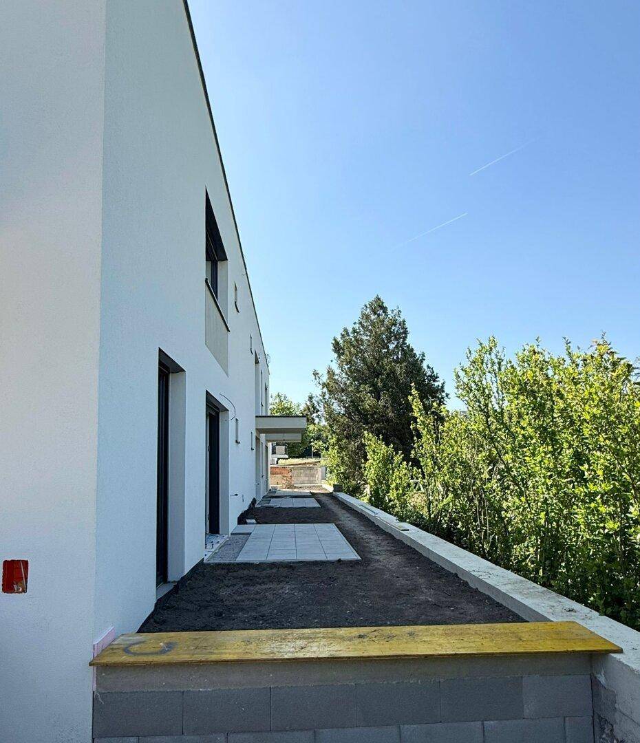 Seitenansicht des Neubaus mit einer frisch angelegten Terrasse und angrenzendem Grünbereich unter blauem Himmel.