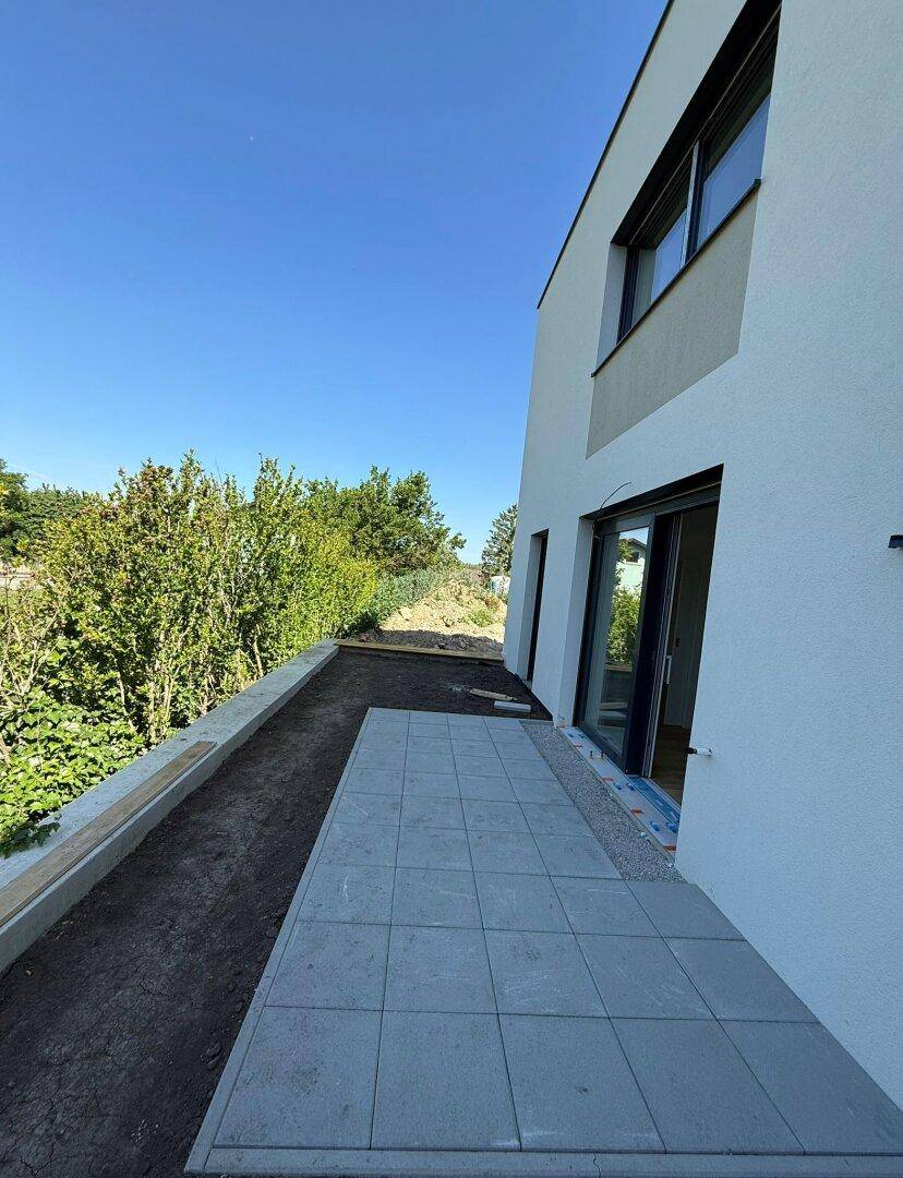 Neu gepflasterte Terrasse am modernen Gebäude, umgeben von üppigem Grün und unter strahlend blauem Himmel.