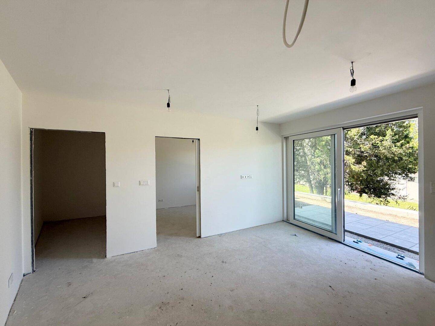 Geräumiger, unfertiger Raum mit Betonboden und großem Schiebefenster, das viel Tageslicht hereinlässt.