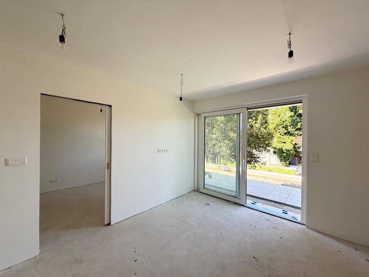 Unfertiger Raum mit Betonboden, weißen Wänden und großem Schiebefenster zum Außenbereich.
