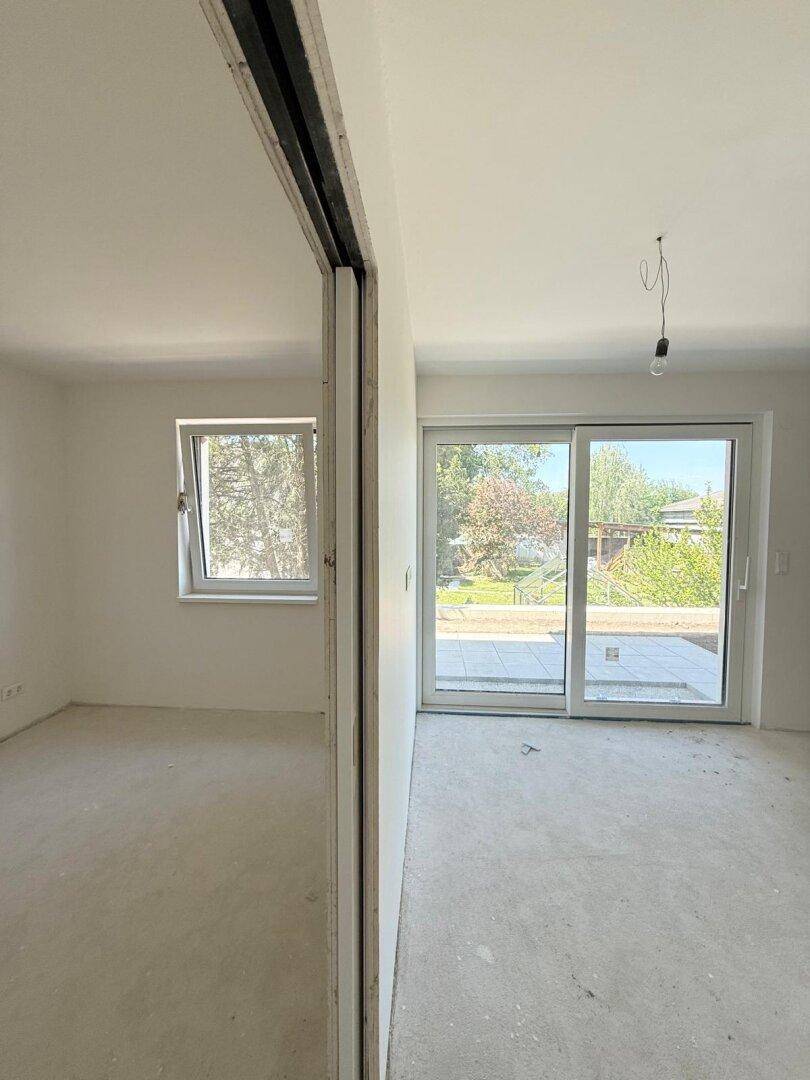 Helles Zimmer im Rohbau mit Betonboden und großen Fenstern, die den Blick ins Grüne freigeben.