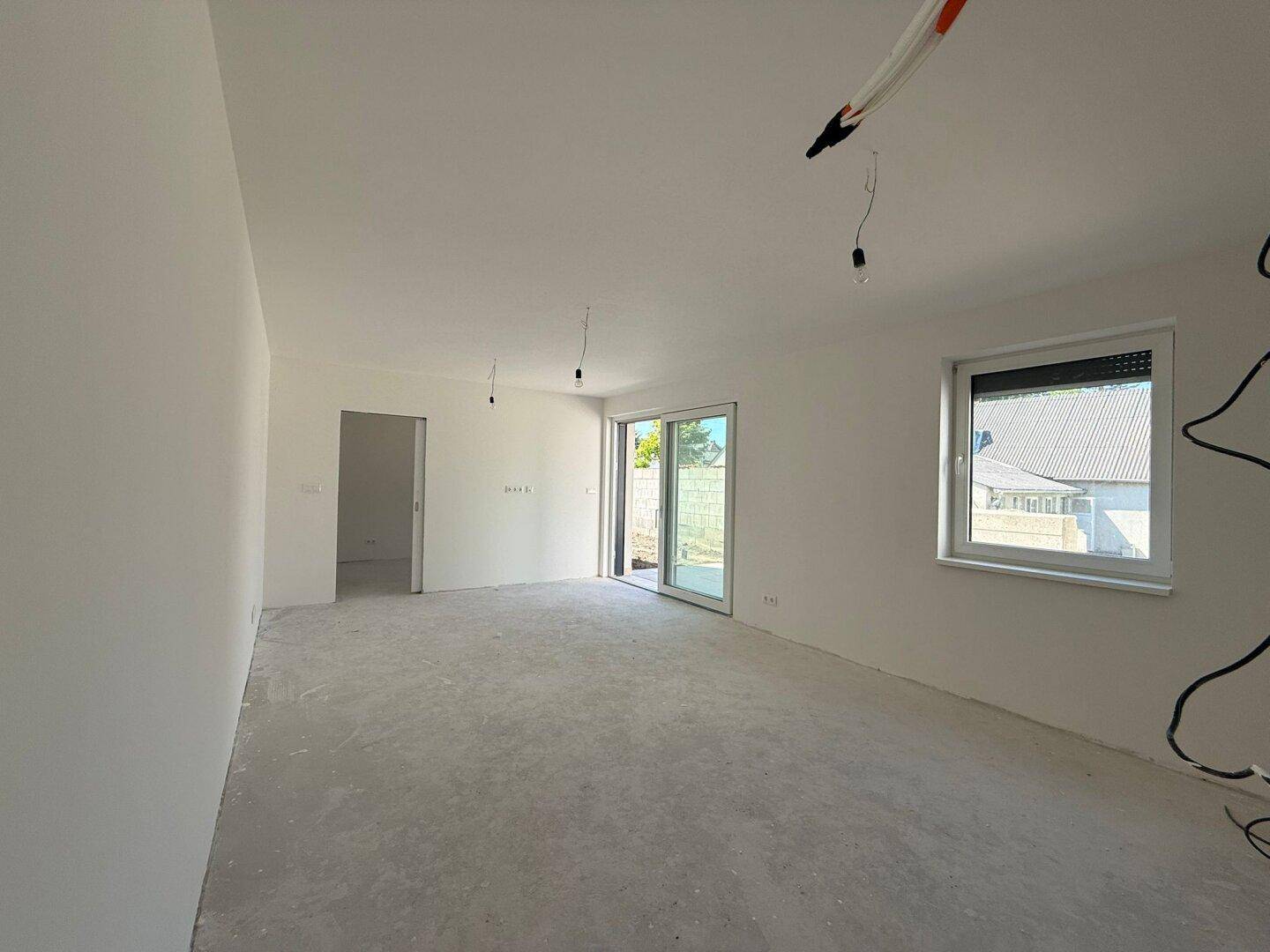 Heller Rohbau-Raum mit Betonboden, großen Fenstern und vorbereiteten Elektroinstallationen.
