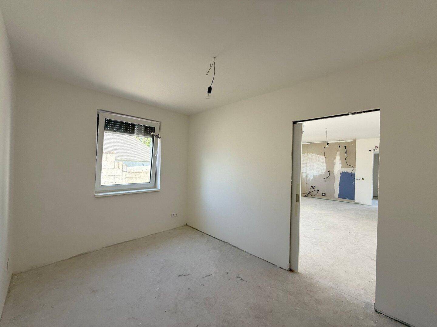 Unfertiger Raum mit Betonboden und Fenster, der durch eine Schiebetür mit einem größeren Bereich verbunden ist.