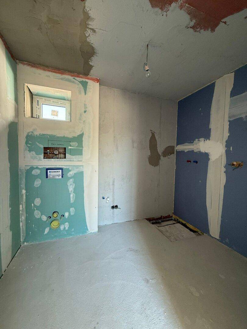 Badezimmer im Rohbau mit vorbereiteten Installationen für WC und Waschbecken sowie einem Fenster.