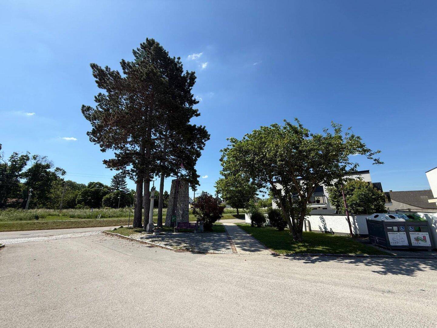 Weitläufige Außenansicht mit Bäumen, einem Denkmal und Gebäuden im Hintergrund unter blauem Himmel.