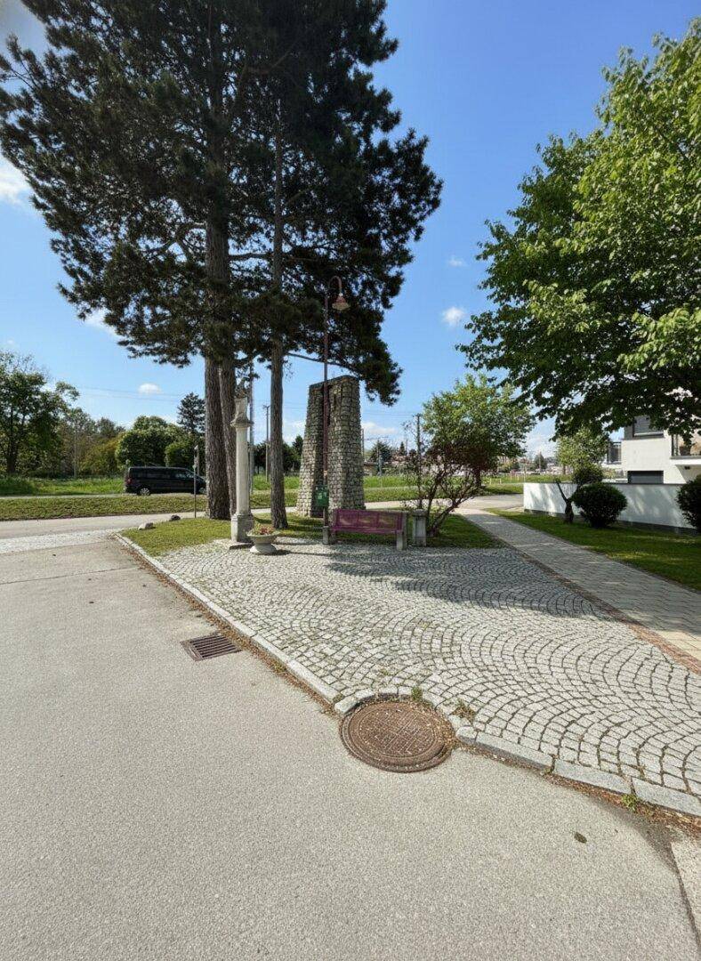 Kleiner öffentlicher Platz mit Kopfsteinpflaster, Bäumen und einem Denkmal unter strahlendem Sonnenschein.