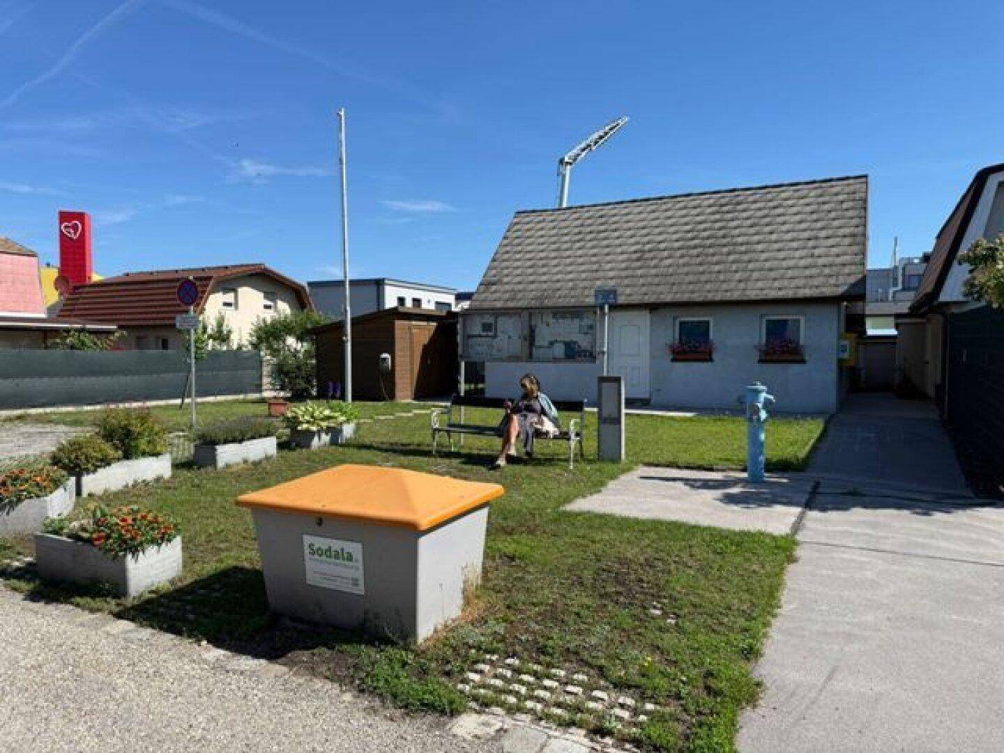 Kleines Haus mit Rasenfläche und Sitzbank, umgeben von Blumenbeeten unter blauem Himmel.