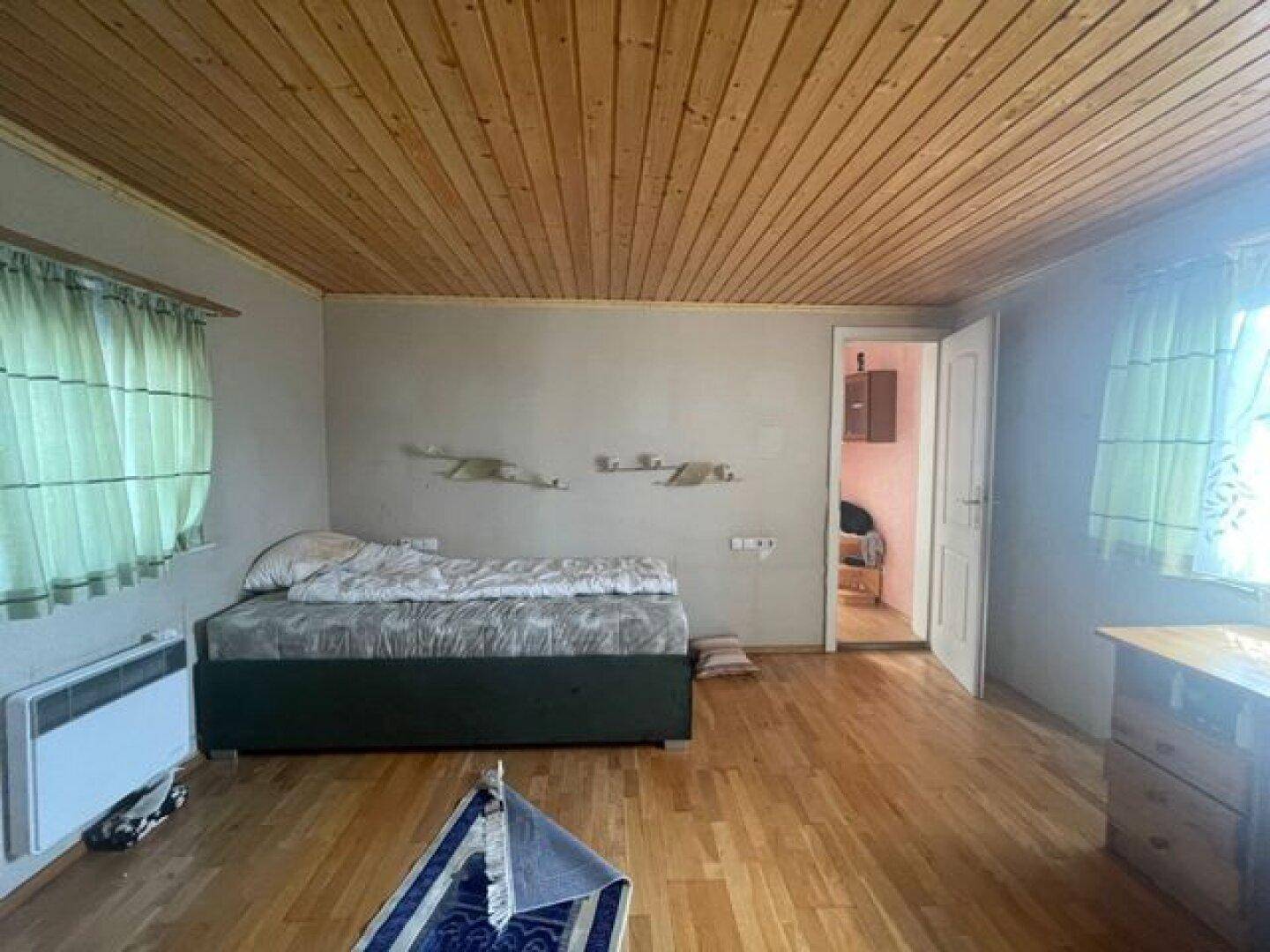 Schlafzimmer mit Holzbalkendecke, Holzboden und einem Bett, hell durch zwei Fenster.