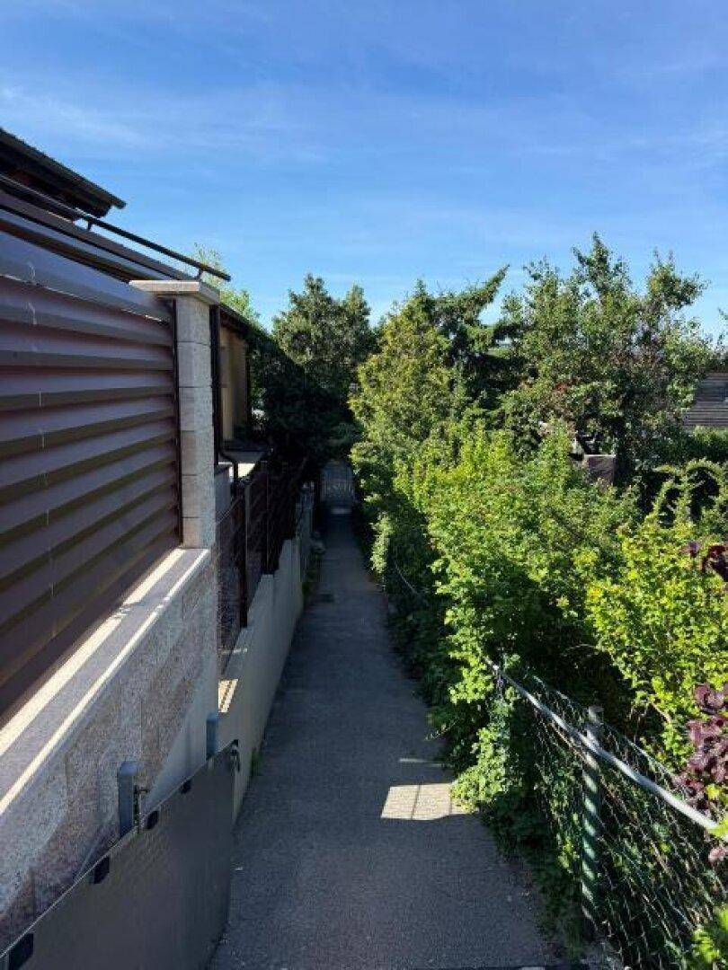 Schmaler Weg zwischen Gebäuden, umgeben von hohen Hecken und Zäunen unter blauem Himmel.