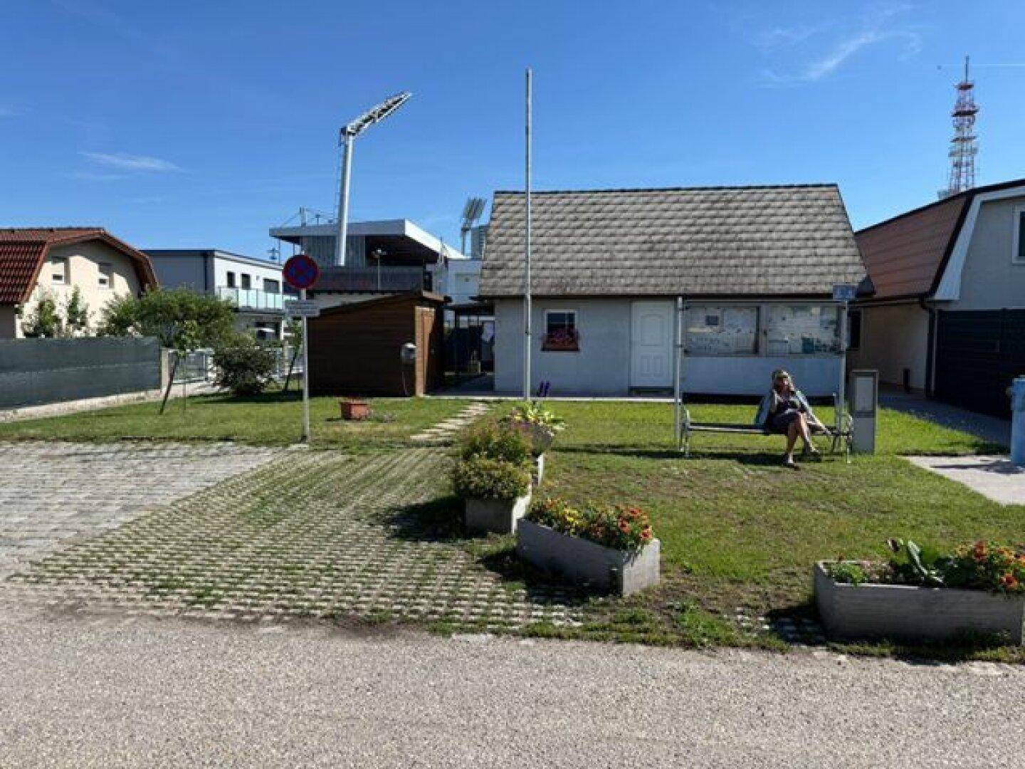 Übersicht über ein kleines Haus mit Rasenfläche, Blumenbeeten und einer Sitzbank im Freien.