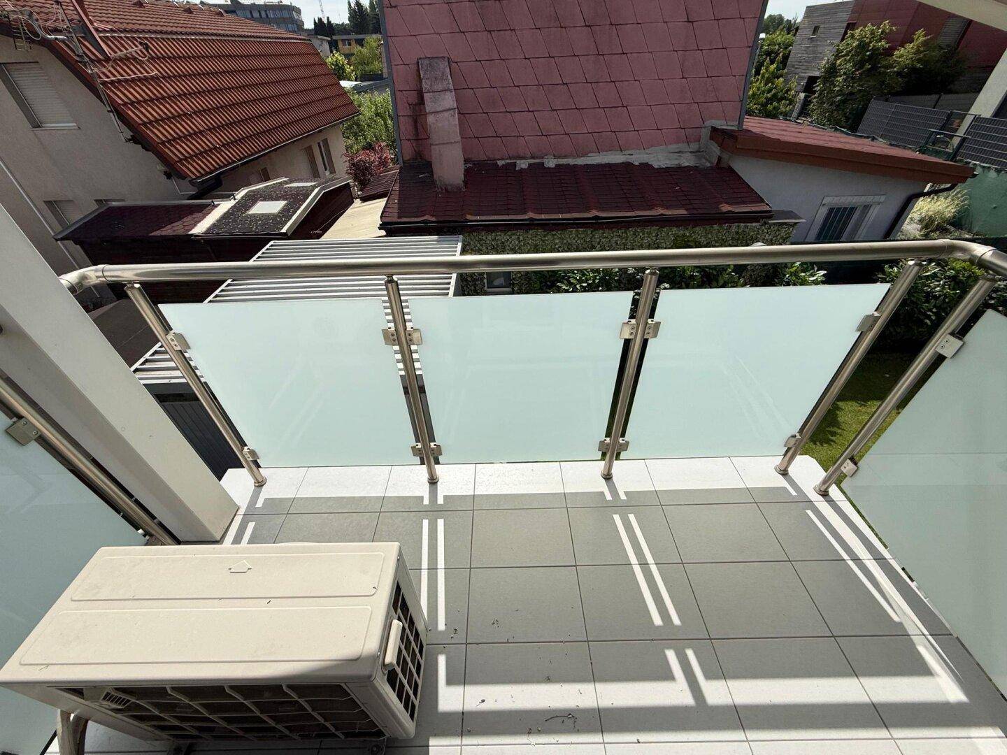 Balkon mit Fliesenboden, Glasgeländer und Blick auf die umliegenden Dächer.
