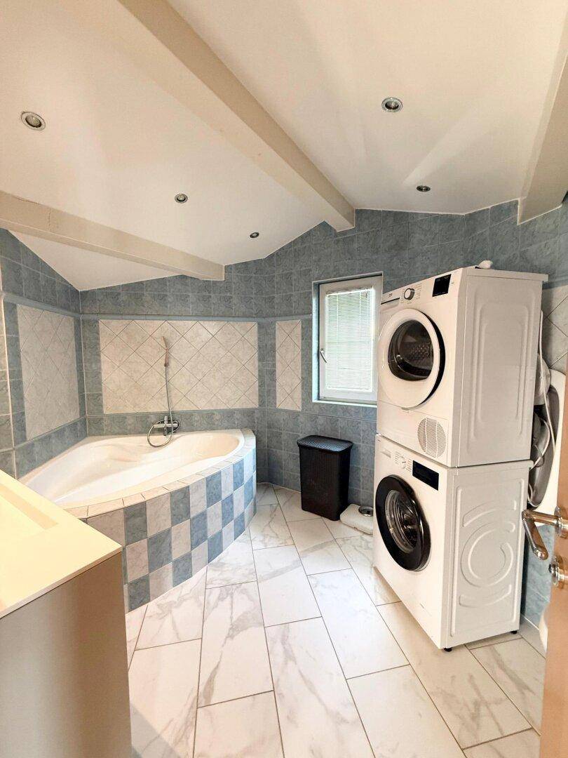 Badezimmer mit blauen Fliesen, Eckbadewanne und praktischem Waschmaschinen-Trockner-Turm.