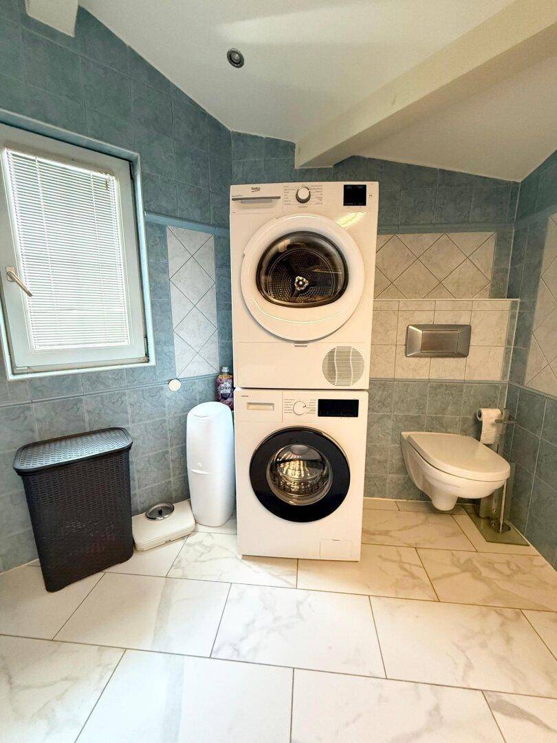 Funktionales Badezimmer mit Waschmaschine, Trockner, Toilette und Fenster für Tageslicht.