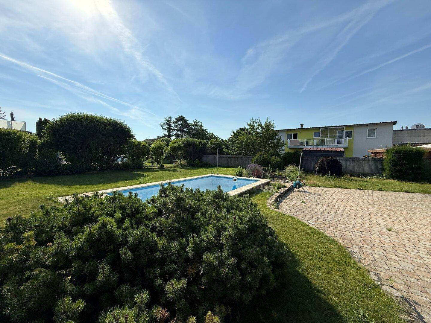 Weitläufiger Gartenbereich mit Swimmingpool und gepflasterter Fläche unter blauem Himmel.