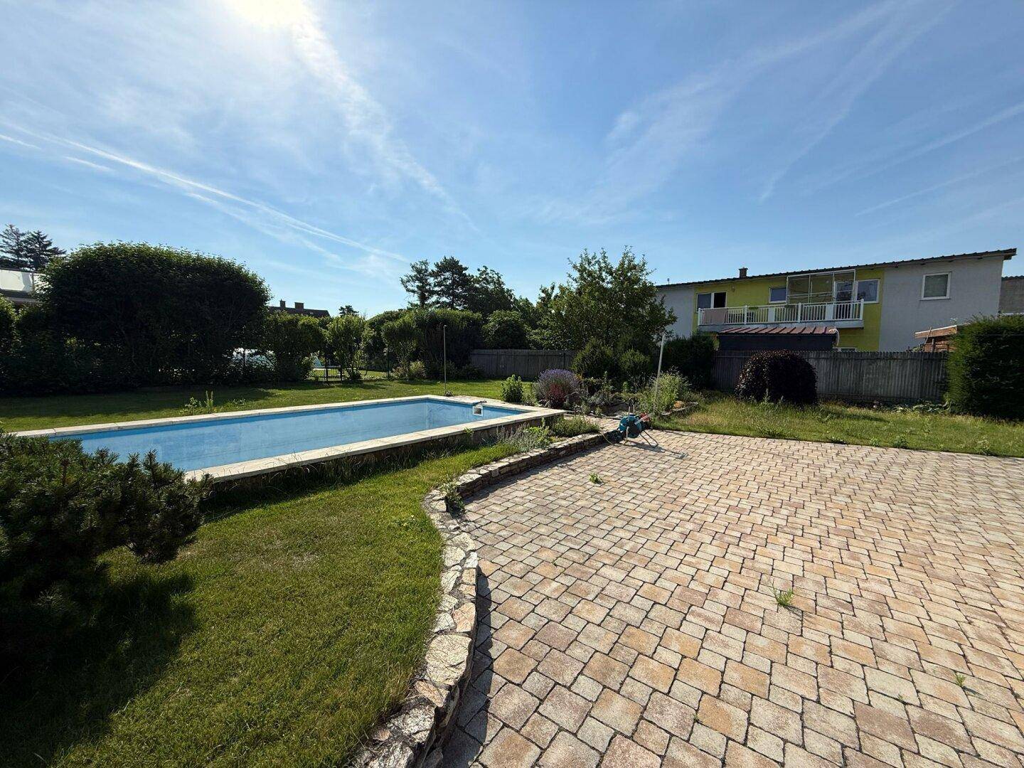 Sonniger Garten mit rechteckigem Swimmingpool und großzügiger gepflasterter Terrasse.