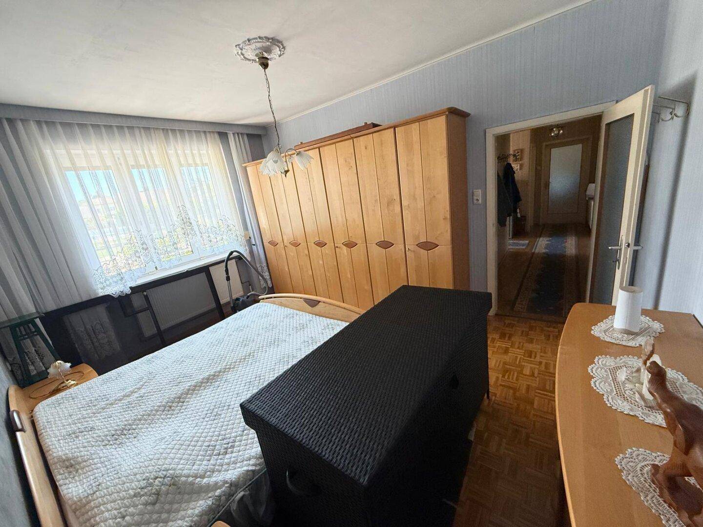 Schlafzimmer mit Doppelbett, großem Holzschrank und Fenster mit Vorhängen.