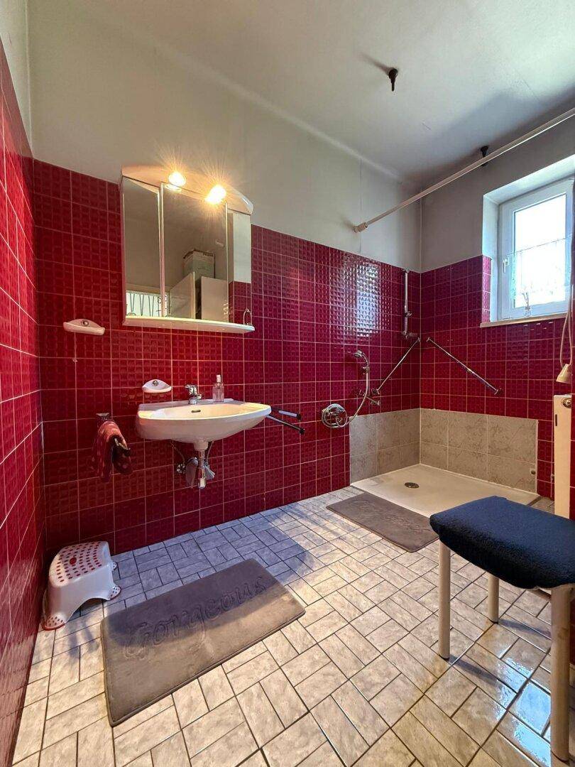Badezimmer mit roten Fliesen, Waschbecken und barrierefreier Dusche mit Haltegriffen.