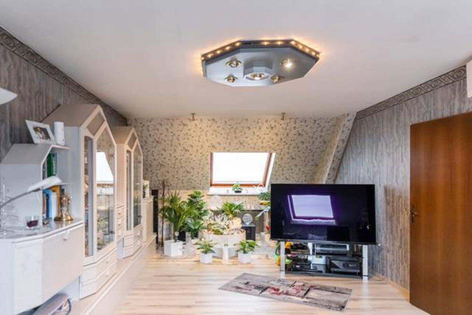Helles Wohnzimmer in der Dachgeschosswohnung mit Fernseher, Vitrinen und Pflanzen.