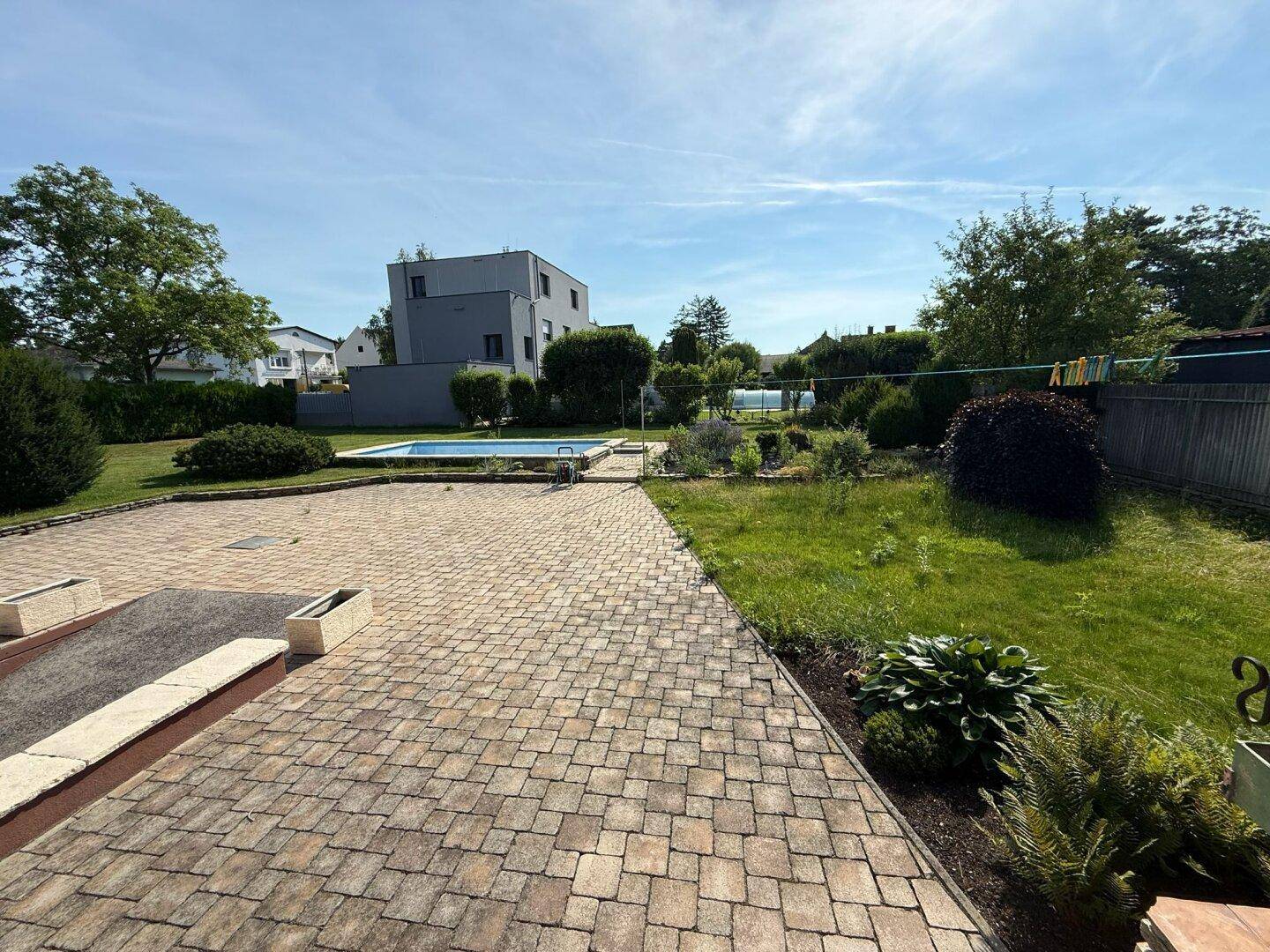 Weitläufiger Gartenbereich mit gepflasterter Fläche und Pool im Hintergrund, umgeben von Grün.