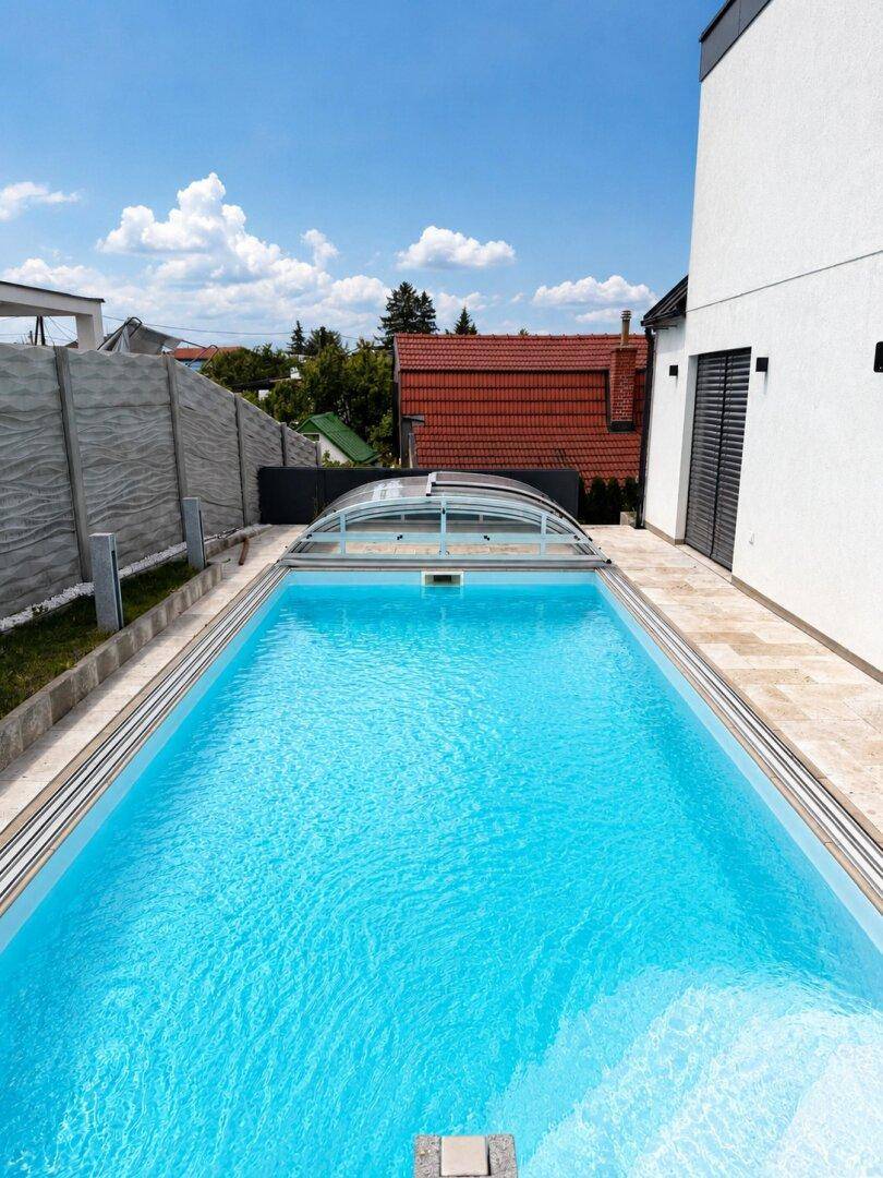 Langer, rechteckiger Pool mit Schiebeüberdachung und gepflastertem Bereich neben dem Haus.